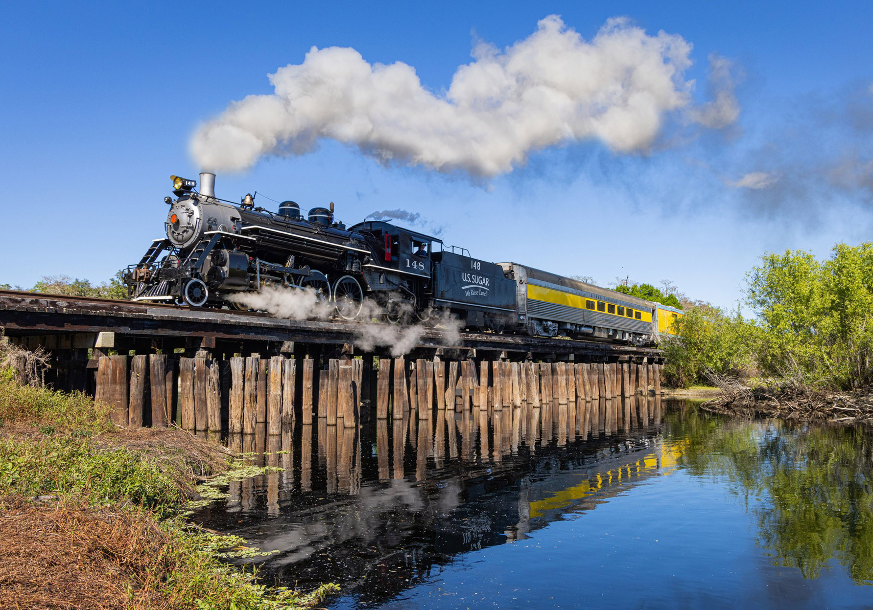 Steam train crossing a wooden bridge over a reflective river on a sunny day.