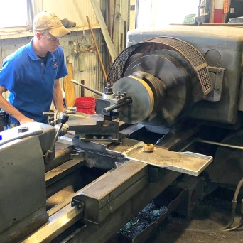 Person in a blue shirt operating an industrial metal lathe in a workshop.