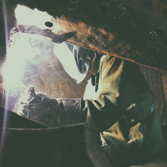 Person welding with sparks and protective gear in dim light.