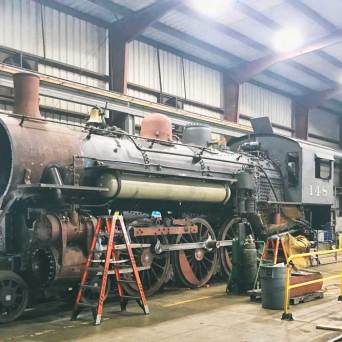 Old steam locomotive in a large indoor workshop with ladders and tools around.