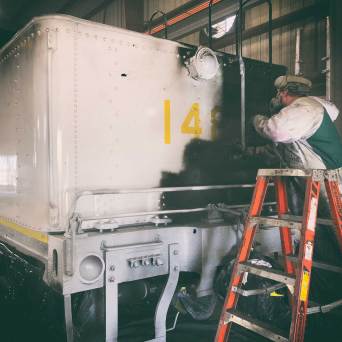 Person spray-painting a large vehicle part on a ladder.
