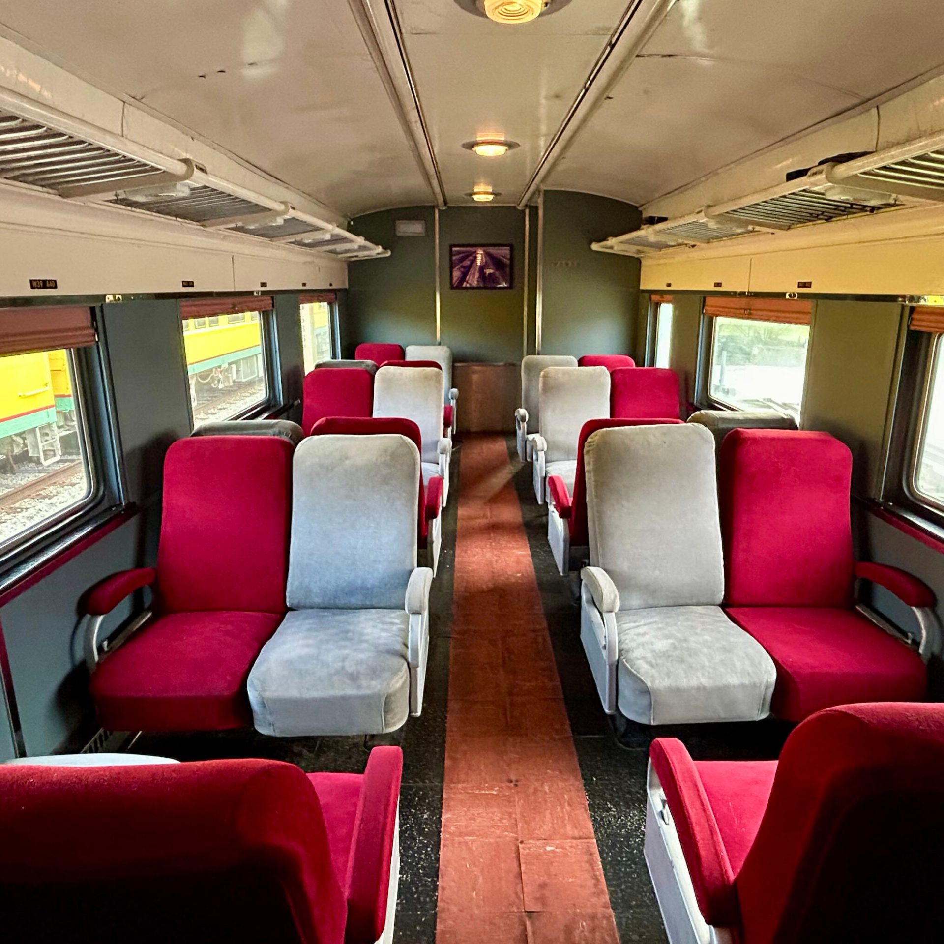 Empty train cabin with red and grey seats, overhead luggage racks, and windows.
