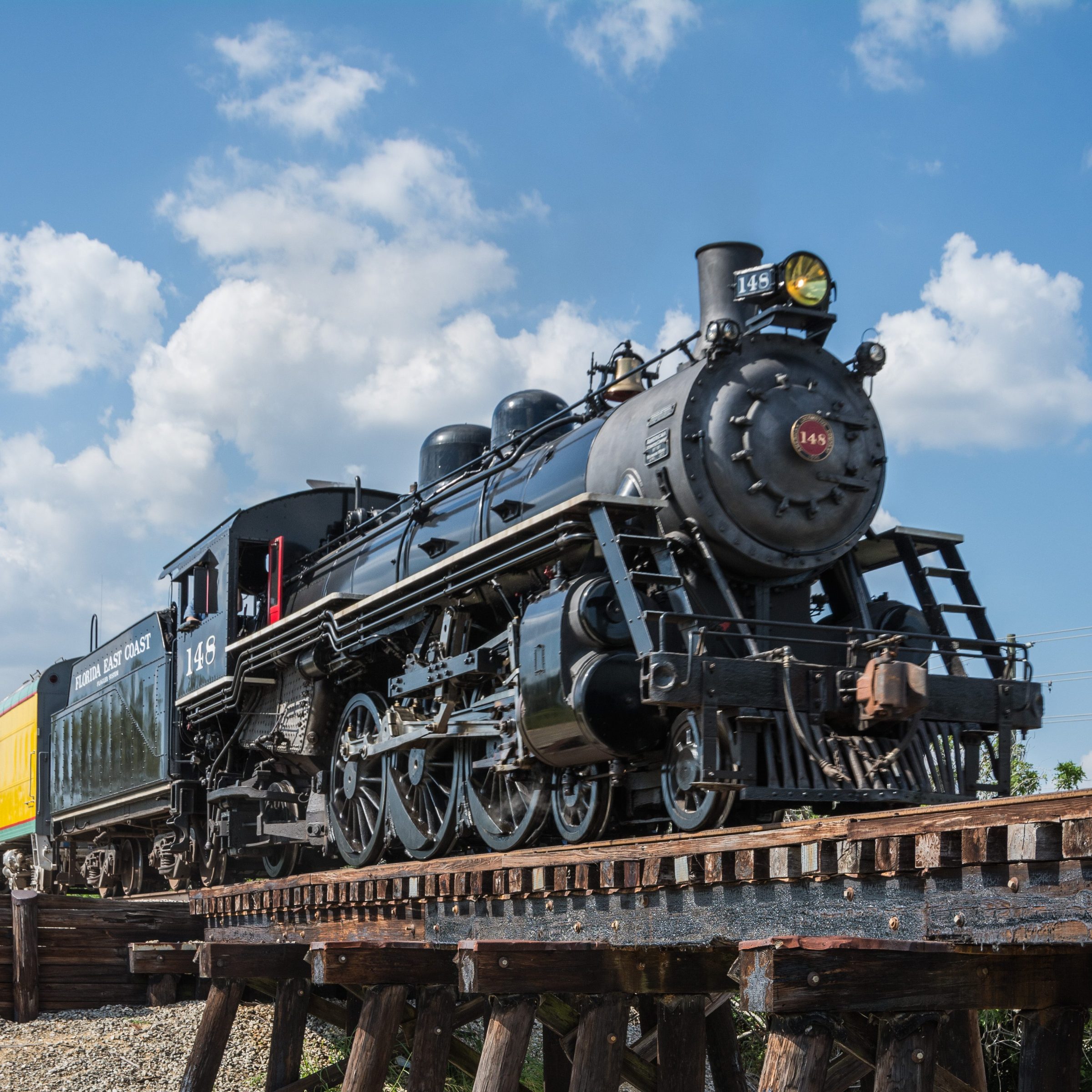 Steam locomotive on wooden trestle with vintage car below, under a blue sky.