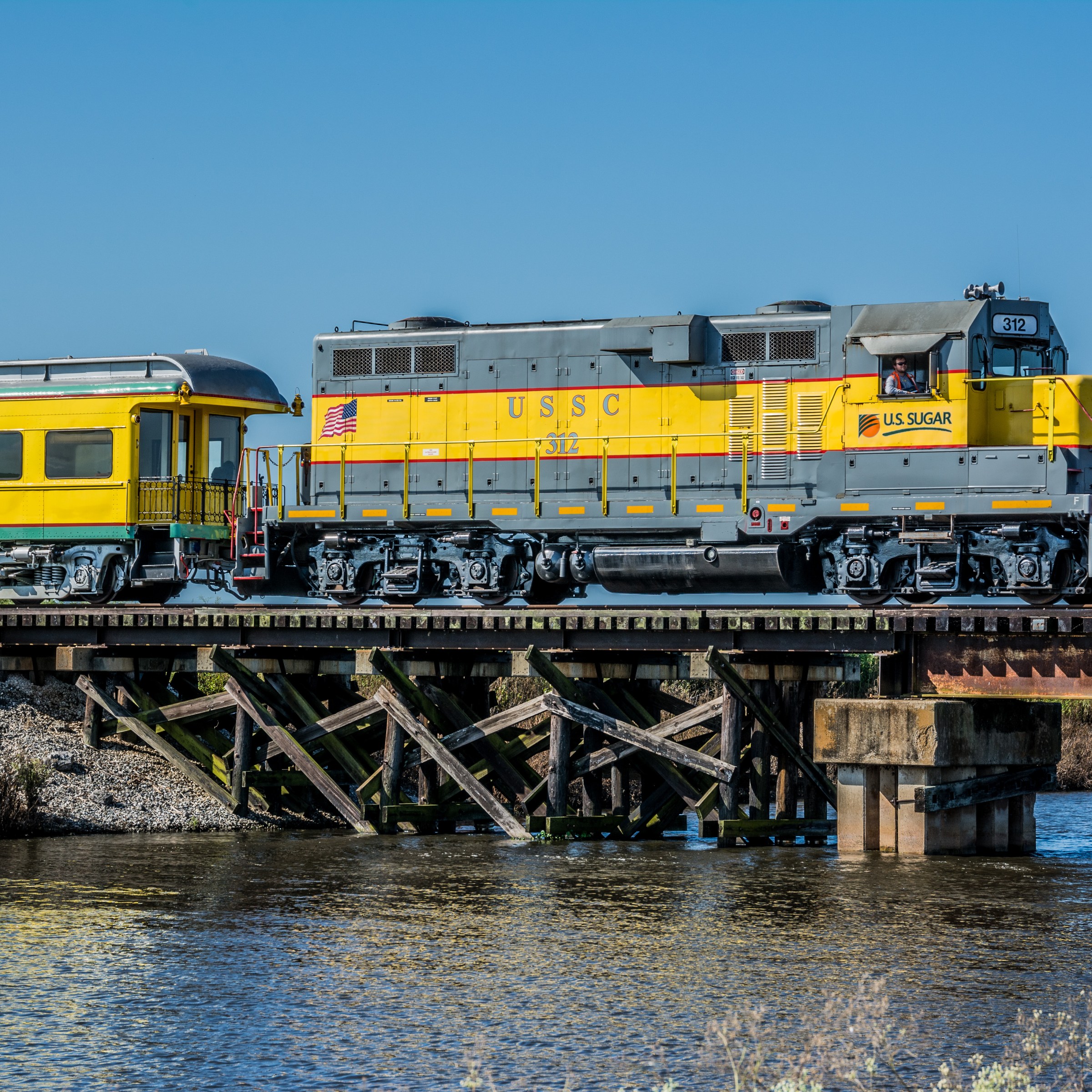Yellow train crossing a wooden bridge over a calm river.