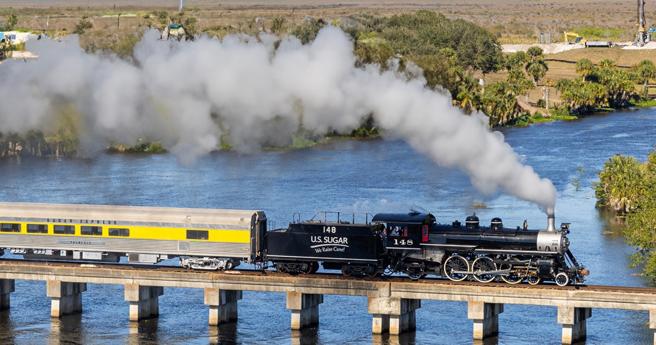 Aerial view of U.S. Sugar steam locomotive pulling Sugar Express passenger car across bridge over Florida waterway
