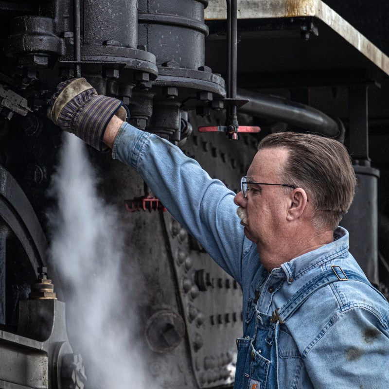 Man in denim overalls adjusts steam valve on a vintage train engine.