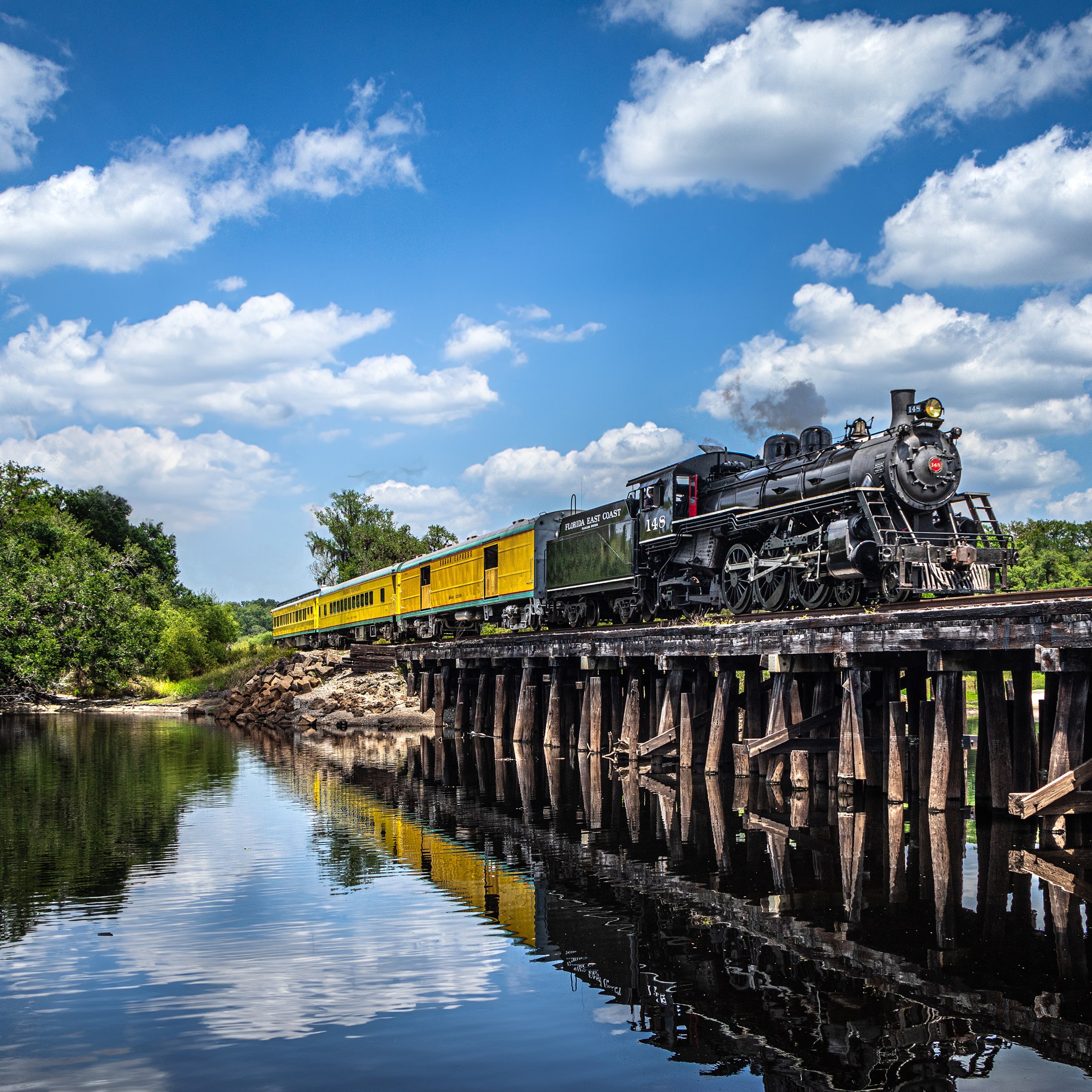 Steam train crossing a wooden bridge over a river, surrounded by trees and blue sky with clouds.