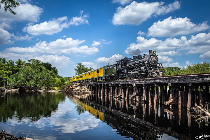 Steam train crossing a wooden bridge over a river, surrounded by trees and blue sky with clouds.