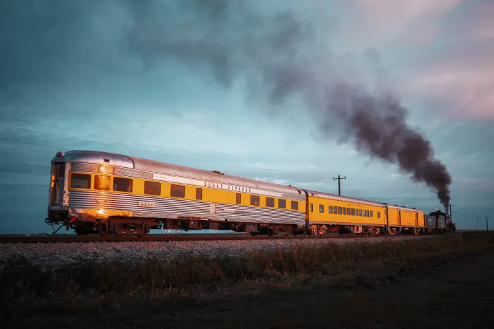 Vintage yellow train emitting smoke under a cloudy sky.