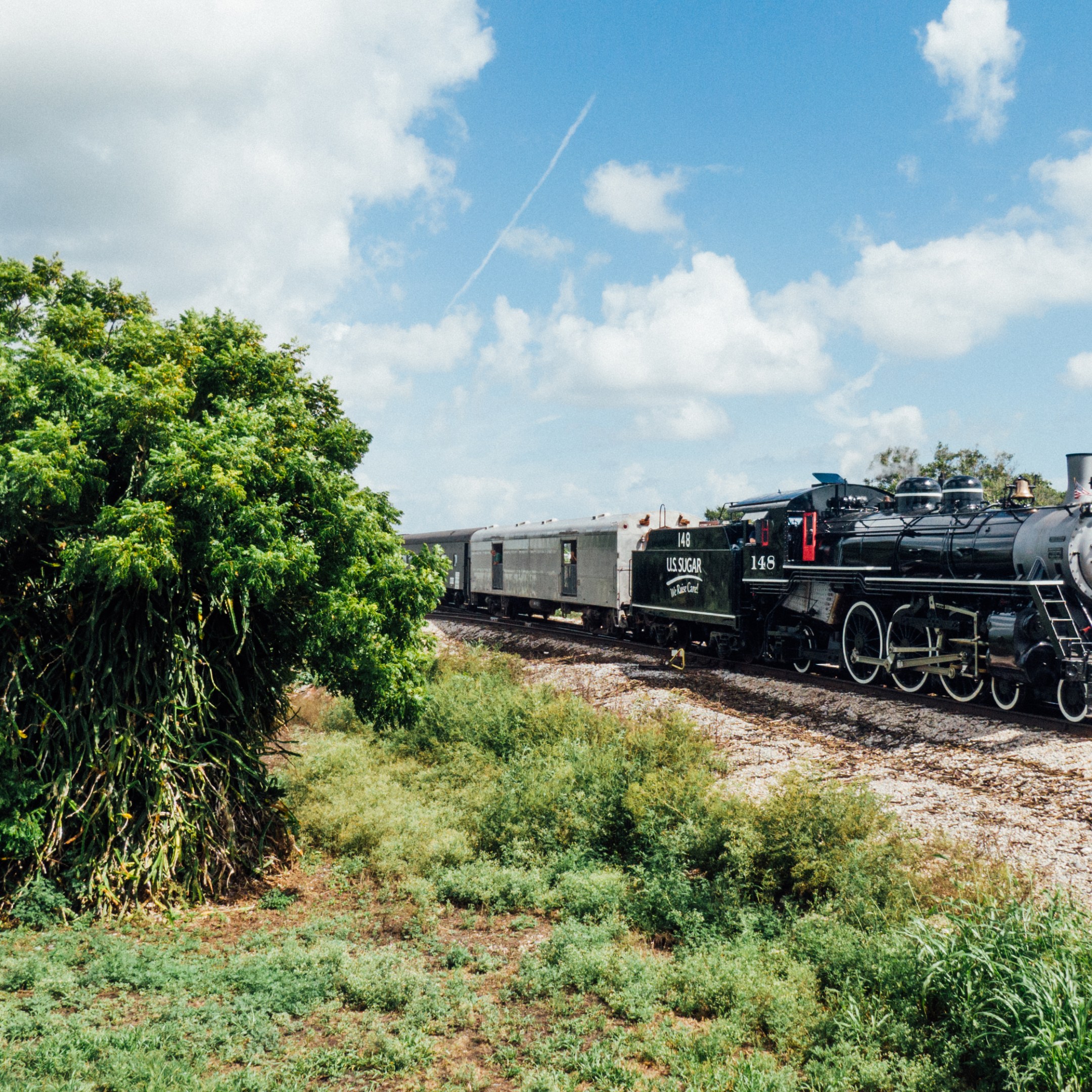 Steam train on tracks surrounded by grass and trees under a cloudy sky.