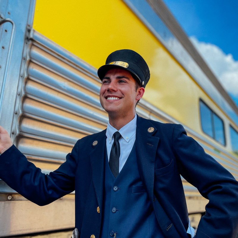 Train conductor smiling beside a yellow train under blue sky.