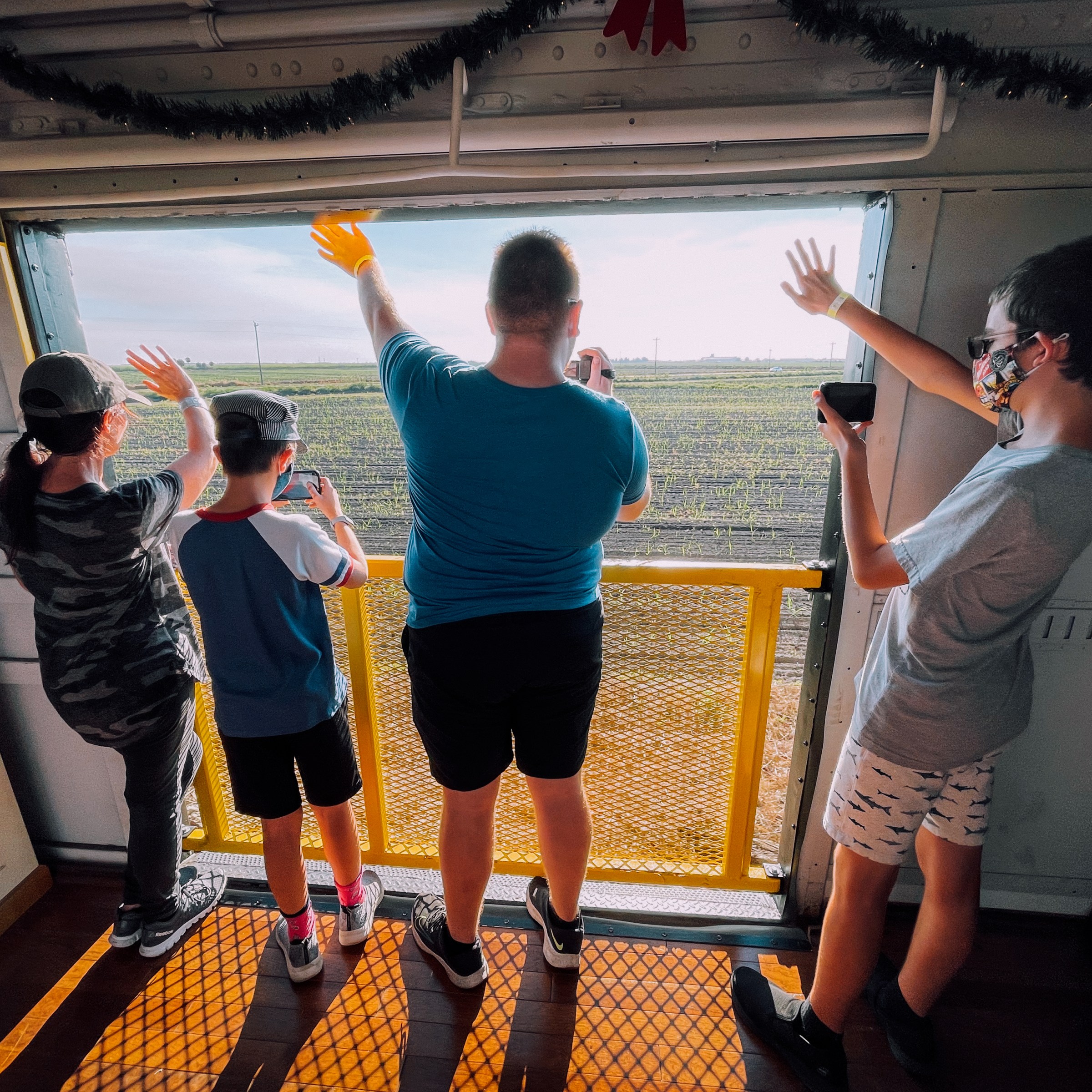 Four people wave from a train, looking out at scenic countryside.
