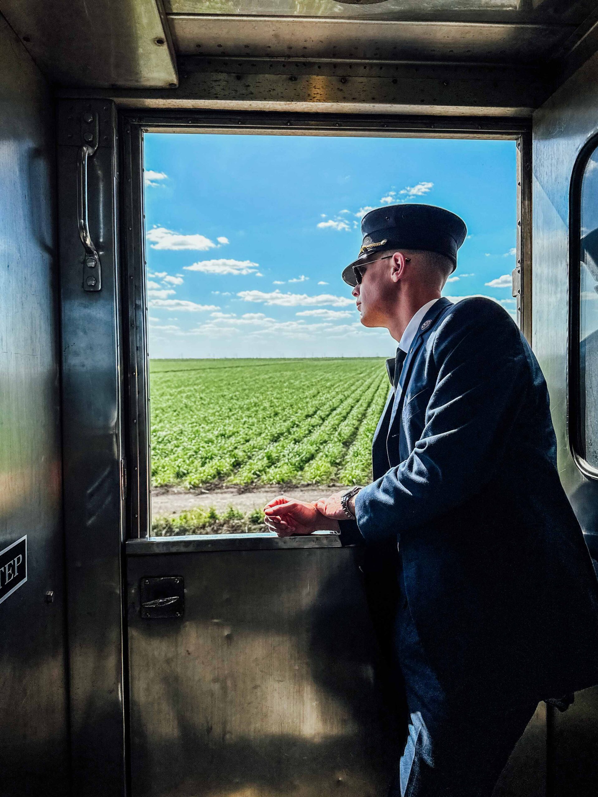Train conductor in uniform looking out open doorway of Sugar Express railcar across green agricultural fields