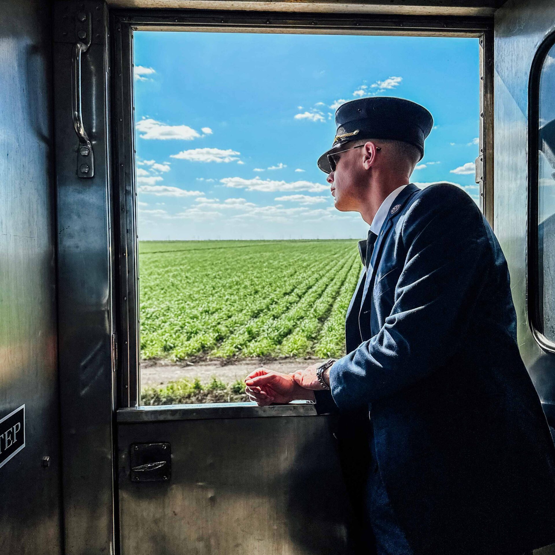 Person in a blue uniform looking out a train window at a green field under a blue sky.