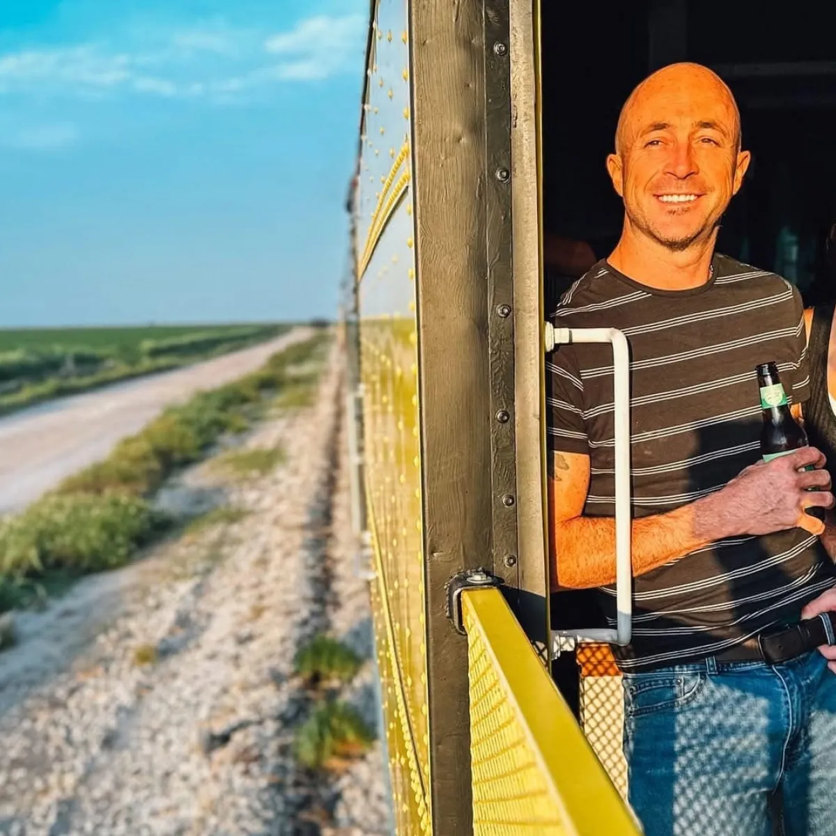 Two people smiling, standing on a moving train, with open landscape view.