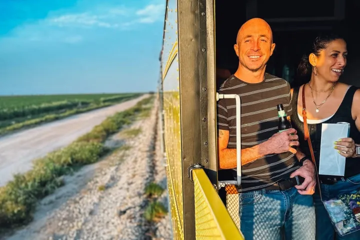 Two people smiling, standing on a moving train, with open landscape view.