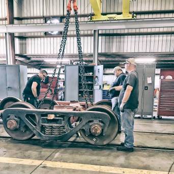 Workers inspecting a train wheelset in an industrial workshop with chains overhead.