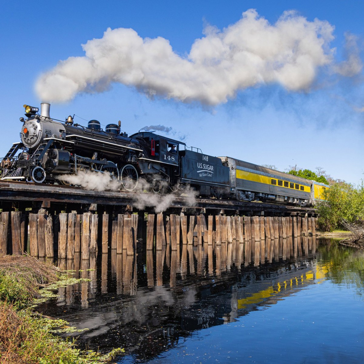 Steam locomotive and yellow train car cross wooden bridge over water.