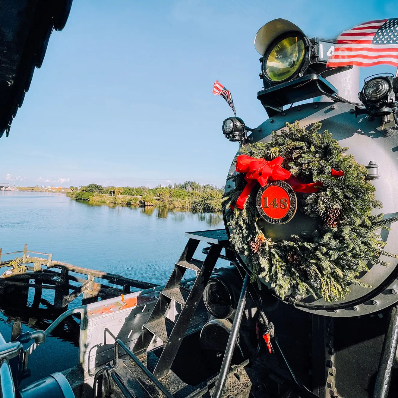 Steam train with wreath and U.S. flags near a river in a sunny landscape.