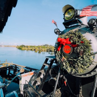 Steam train with wreath and U.S. flags near a river in a sunny landscape.