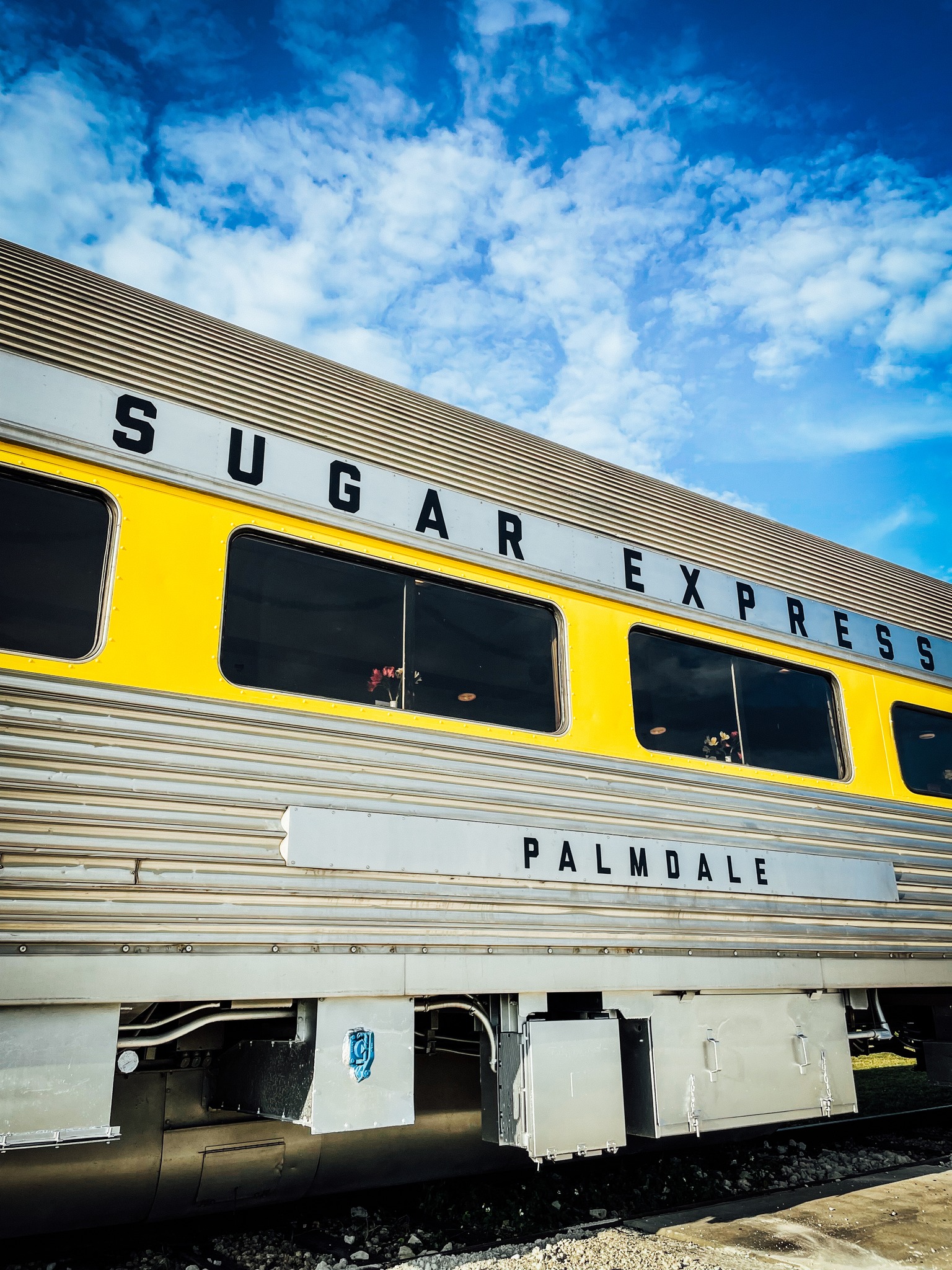 Exterior side view of bright yellow Sugar Express passenger railcar labeled Palmdale under blue sky