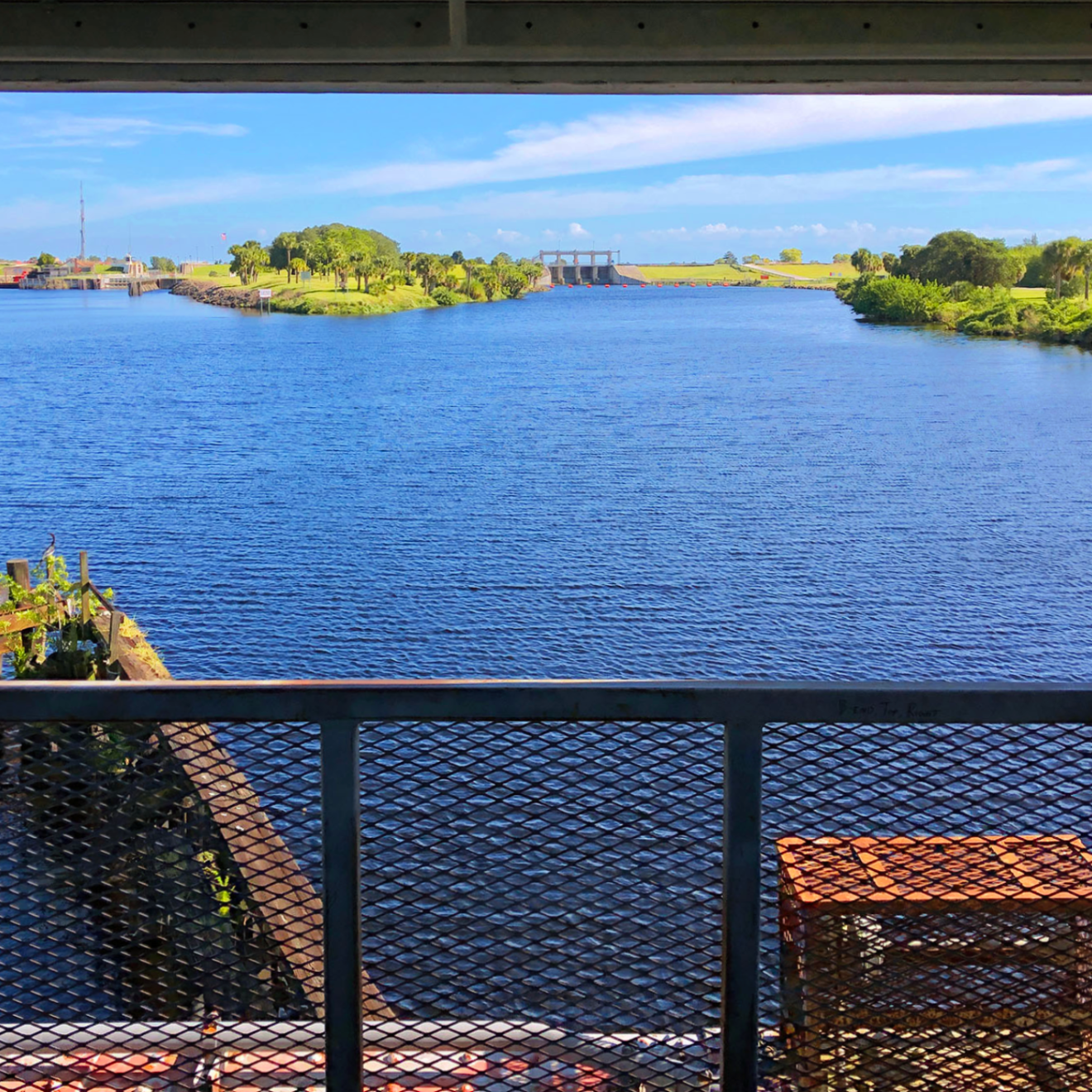 View through a fence of a river with greenery and a bridge in the distance under a blue sky.