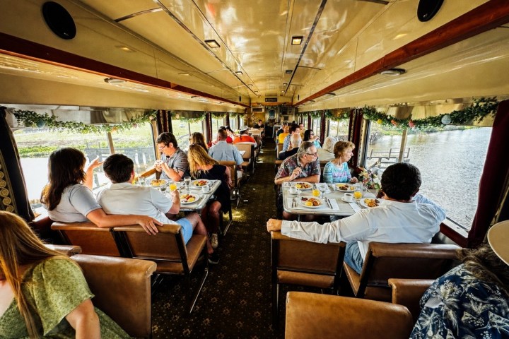 Passengers dining in a vintage train car with large windows and tables set with food.